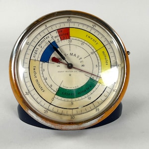 May include: A round Merit Watch Co. Inc. Sport-Master timer with a metallic orange outer rim and a dark blue base. The off-white face features black text, a red, yellow, blue, and green color-coded dial, and black hands with a red pointer.