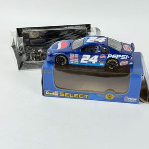 May include: A blue and white die-cast model race car with the number 24 on the roof and sides, featuring Pepsi and Fritos logos. The car is displayed on a blue box with the Revell Select logo. A clear plastic case is in the background.