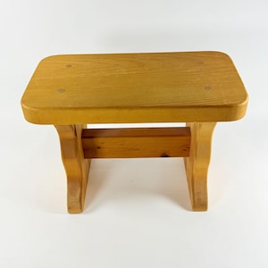 Peut inclure: Un tabouret rectangulaire en bois avec une finition brun clair, doté d'un dessus plat et lisse avec quatre bouchons en bois circulaires visibles et deux supports latéraux incurvés reliés par une entretoise horizontale au centre.