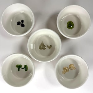 May include: Five small, white ceramic bowls. Each bowl features a different food-themed design: olives, basil, garlic, broccoli, and mushrooms. The bowls are arranged in a circular pattern, perfect for serving dips or small portions.