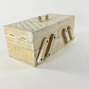 May include: A rectangular wooden sewing box with a light, whitewashed finish. The lid features a carved geometric lattice pattern and two small wooden knobs. The box includes a tiered, accordion-style folding mechanism with hinged wooden arms.