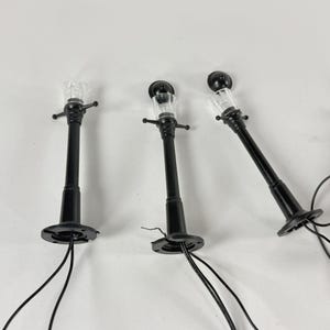 May include: Three miniature black street lamps with clear, textured light covers. Each lamp has a round base and a small, decorative top. Black wires extend from the base of each lamp.
