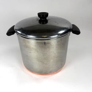 May include: A stainless steel stockpot with a matching lid, featuring two black side handles and a black knob on the lid. The cylindrical pot has a polished metallic finish on the sides and a visible copper-coloured base at the bottom.