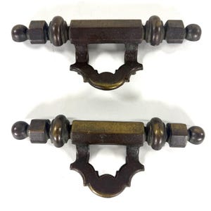 May include: Two antique bronze drawer pulls. Each pull features a rectangular center with decorative end pieces and a curved, ornate handle. The pulls are designed for furniture and have a vintage aesthetic.