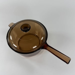 May include: A round, transparent amber-colored glass saucepan with a matching glass lid. The cookware features a long, integrated glass handle extending from one side and a small knob on the center of the lid for easy removal and handling.