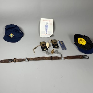 May include: A collection of vintage scout memorabilia, including two navy blue caps with yellow and gold accents, a brown leather belt, epaulettes, and a document titled "Inspection Report". The items are arranged on a gray surface.