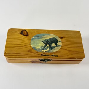 May include: A rectangular wooden box with a natural grain finish and a dark knot on the left side. The lid features an oval illustration of a black bear catching a fish in a stream, with the text Jackman, Maine printed in black below the image.