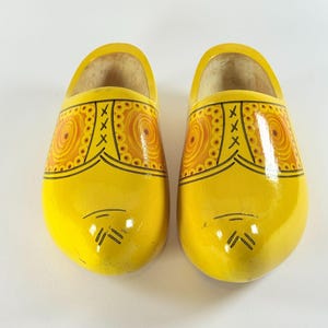 May include: A pair of bright yellow wooden clogs featuring a glossy finish and decorative orange circular patterns on the top. The shoes are adorned with black line details that mimic the appearance of laces and stitching across the front.
