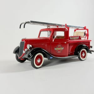 May include: A red and black die-cast model fire truck featuring a silver ladder mounted on the roof, a white hose reel in the truck bed, and a circular Fire Dept emblem on the door. The vehicle has white-walled tires and a classic truck design.