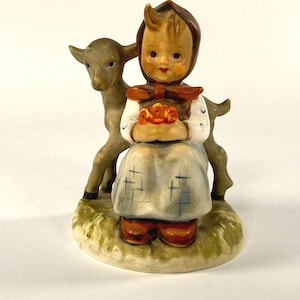 May include: A ceramic figurine of a girl in a bonnet and dress, holding a basket of flowers, with a lamb standing behind her. The figurine has a painted finish with brown, blue, white, and green colors. The base is oval-shaped and painted green.