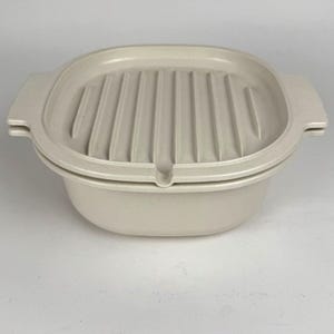 May include: A light beige oval-shaped casserole dish with a ridged lid. The dish has two handles and a small notch on the side. The lid has a series of parallel ridges.