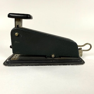 May include: A vintage stapler in a dark green hue, featuring a black handle and a silver metal lever. The stapler has a rectangular base and a classic design, showcasing its age and use.