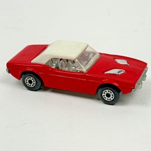 May include: A red die-cast toy car featuring a white roof, clear plastic windows, and silver-colored interior seats. The vehicle has black wheels with silver rims and two silver-colored hood vents on the front hood of the car body.