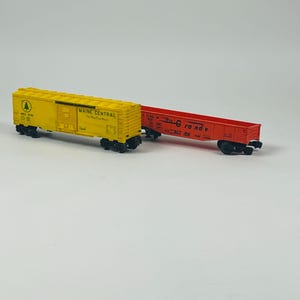 May include: Two model train cars. One is yellow with black wheels and the words "MAINE CENTRAL" printed on the side. The other is red with black wheels and the words "Rio Grande" printed on the side. Both are on a white surface.