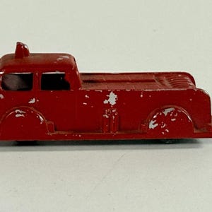 May include: A vintage red toy fire truck with visible wear and tear. The toy has a cab with two windows, a small siren on top, and a flatbed. The paint is chipped, giving it a rustic appearance. A classic collectible.
