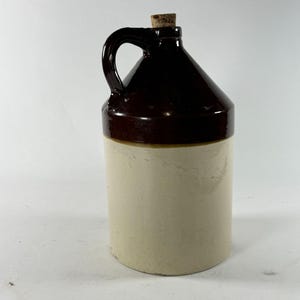 May include: A cylindrical stoneware jug featuring a two-tone design with a glossy dark brown glazed upper section and a matte cream-colored lower body. The jug includes a single side handle, a narrow neck, and a small cork stopper at the top.