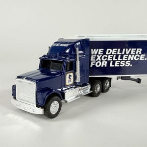 May include: A navy blue U.S. Mail semi-truck toy with a white trailer. The truck has silver accents and the words "WE DELIVER EXCELLENCE. FOR LESS." are printed on the trailer. The words "U.S. MAIL" are printed on the cab.