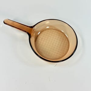 May include: A vintage amber-colored glass skillet with a handle. The pan has a round cooking surface and a handle extending from the side. The skillet is made of transparent glass, allowing visibility of the interior.