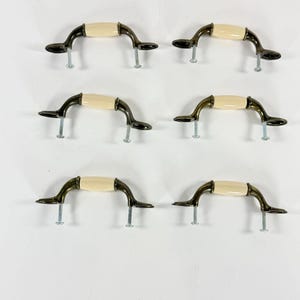May include: Six antique-style cabinet handles. Each handle has a cream-colored center and dark bronze-colored curved arms. The handles are shown with the screws for installation.