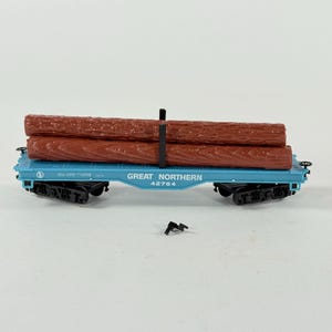 May include: A blue model train flatcar carrying two brown textured plastic logs secured by a black stake. The side of the car features the text GREAT NORTHERN 42764. A small detached black plastic component lies on the surface below the train car.