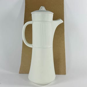 May include: A white ceramic coffee pot with a lid and a handle. The pot has a long, slender body and a spout for pouring. The body of the pot has vertical ribbed detailing. The handle is curved and attached to the side of the pot.