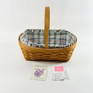 May include: An oval woven wood basket with a single upright handle and a blue, red, and white plaid fabric liner. Included are two paper inserts, one labelled Longaberger Baskets and the other labelled The Easter Basket with floral graphics.
