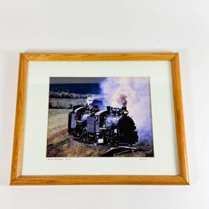 May include: A framed photograph of a black steam locomotive travelling through a grassy landscape, emitting white smoke. The print features the text "107 RE-CREATION" and a signature in the bottom margin, enclosed in a light brown wooden frame.