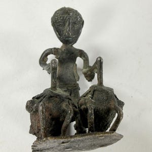 May include: A dark bronze-toned metal figurine depicting a figure standing between two large, rounded, textured containers. The figure has a stylized head and arms extended to hold the handles of the containers, all mounted on a flat base.