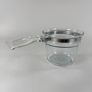 May include: A clear glass saucepan featuring a transparent glass handle and a silver-toned stainless steel band around the upper rim. The metal band is secured with a visible hinge mechanism connecting the handle to the main circular glass vessel.