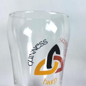 May include: A clear pint glass with the Guinness logo. The logo features the word "Guinness" in black, a black, orange, and red harp symbol, and the word "Harp" below. The glass has a curved shape.
