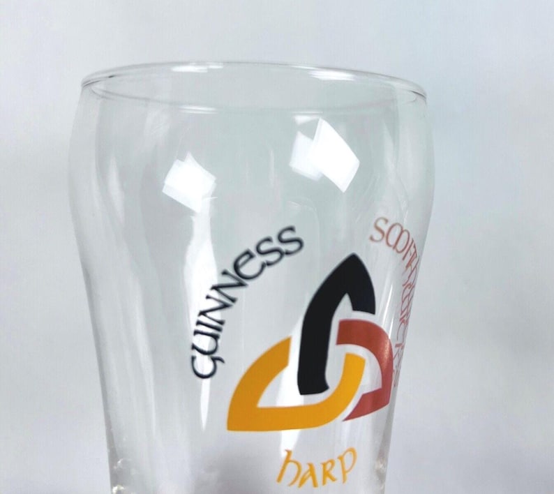 May include: A clear pint glass with the Guinness logo. The logo features the word "Guinness" in black, a black, orange, and red harp symbol, and the word "Harp" below. The glass has a curved shape.