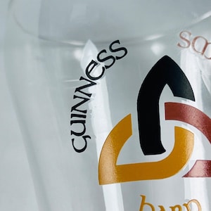 May include: A clear pint glass with the word "Guinness" in black lettering. The glass features a gold, black, and maroon graphic design. The glass is partially filled with a clear liquid.