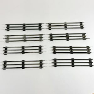 May include: Eight sections of model train track are arranged on a white surface. Each section has three parallel rails connected by evenly spaced black ties. The tracks are made of a dark, metallic material.