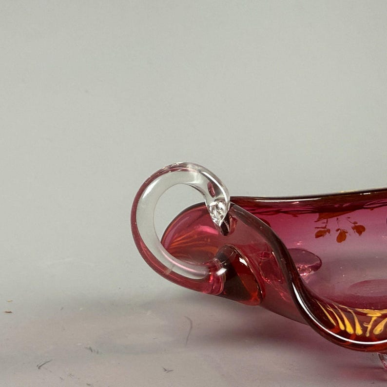 May include: A ruby red glass serving dish with a clear glass handle. The dish has a wavy edge and is decorated with gold painted floral designs.