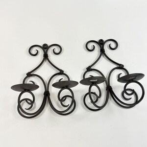 May include: A pair of black metal wall-mounted candle holders featuring symmetrical scrollwork designs. Each piece has two circular platforms for candles and a top loop for hanging, constructed from thin, curved metal rods joined together.