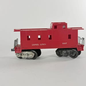 May include: A red Lionel Lines caboose model with black wheels and accents. The model has the number 1007 printed on the side. The caboose has a small roof section with windows and side windows. The words "Lionel Lines" are printed on the side.