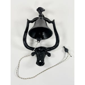 May include: A black cast iron doorbell with a bell-shaped design. The doorbell features a bull's head at the bottom and a rope pull. The bell is approximately 6 inches tall and 5 inches wide.