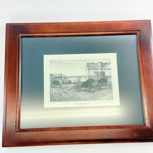 May include: A framed print titled Amish Church Meeting featuring a black and white pencil sketch of horse-drawn carriages parked in front of a building. The artwork is encased in a dark brown wooden frame with a wide black matting border.