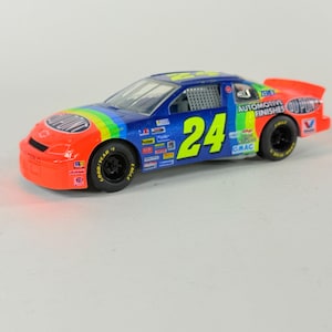 May include: A die-cast model race car, featuring a blue body with rainbow accents and the number 24 in yellow. The car has orange front and rear sections with "DuPont" branding and black wheels. The model is a collectible item.