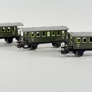 May include: Three model railway carriages. The carriages are dark green with grey roofs and black wheels. Each carriage has multiple rectangular windows with yellow trim. The carriages are arranged in a row.