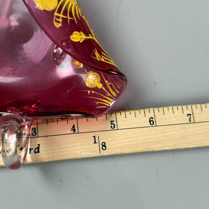 May include: A vintage glass dish with a deep burgundy color and a gold floral design. The dish is approximately 5 inches long and is shown next to a ruler.