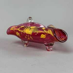 May include: A small, ruby red glass dish with three legs and a handle. The dish is decorated with gold painted floral designs.