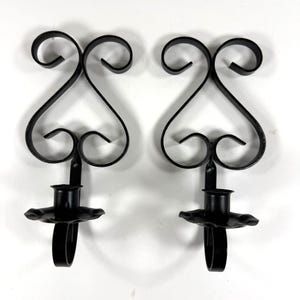 May include: A pair of black metal wall sconces featuring a symmetrical scrollwork design at the top and a circular candle holder base with a scalloped edge. The metal is finished in a solid matte black color, and the structure consists of curved metal strips.