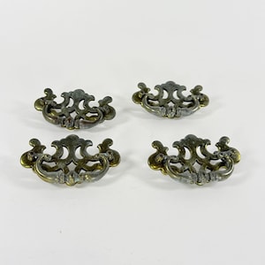 May include: Four antique brass drawer pulls with ornate, curved designs. The pulls have a tarnished, aged appearance, with a mix of gold and gray tones. Each pull features a decorative, symmetrical pattern, suitable for vintage furniture restoration.