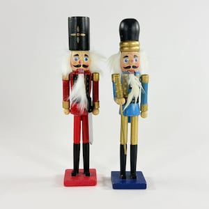 May include: Two wooden nutcrackers, one in red and black with a silver sword, the other in blue and gold. Both have white beards, mustaches, and black hats. They stand on red and blue bases, respectively. A festive holiday decoration.