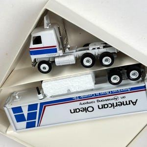 May include: A white and blue toy semi-truck and trailer set. The truck has a white cab with blue and red stripes. The trailer is white with blue geometric shapes and the text "American Olean is Ceramic Tile".