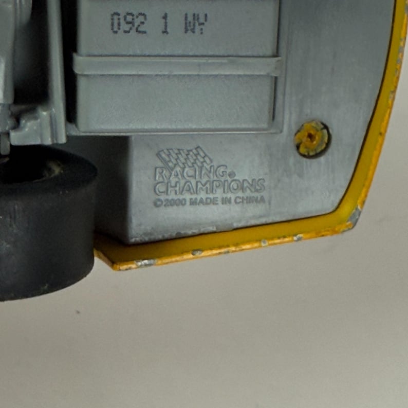 May include: Close-up of the underside of a toy car. The car's base is gray with the text "RACING CHAMPIONS" and "&copy;2000 MADE IN CHINA". The car has a yellow edge and the number "092 1 WY" is visible.