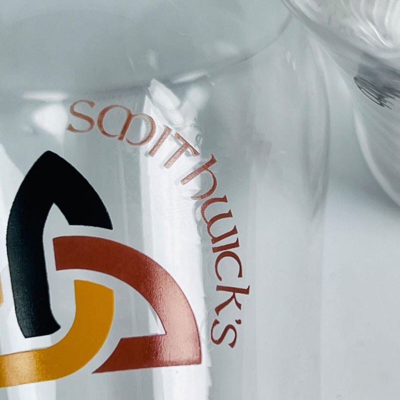 May include: A clear glass with the name "Smithwick's" in reddish-brown lettering. The logo includes a black, brown, and yellow design. The glass is partially visible against a bright background.
