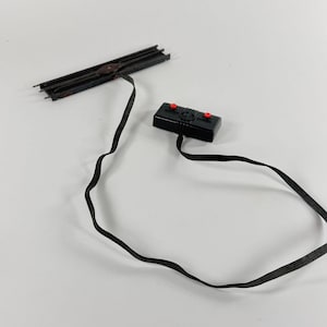 May include: A black toy train track section connected by a black wire to a black control box with two red buttons. The track is dark brown and has a weathered appearance.