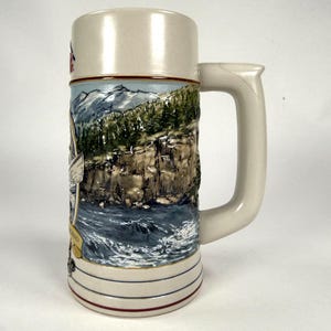 May include: A ceramic beer stein featuring a relief-molded landscape scene of a rocky cliffside, a forest, and a river. The stein has a cream-colored base, a sturdy handle, and a decorative band at the bottom with thin red and blue lines.